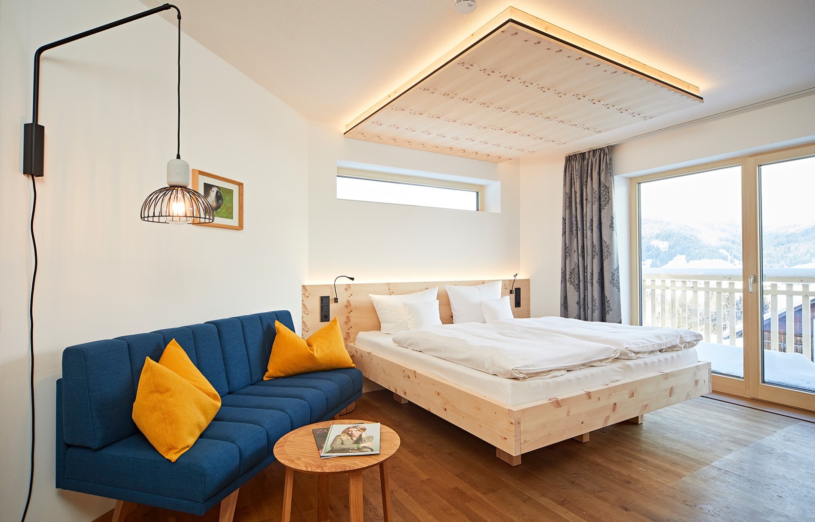 Modernes Hotelzimmer mit blauem Sofa, Beleuchtung, Fernseher und Balkon in hellen Holzfarben im Kleinwalsertal Hotel Oswalda Hus.