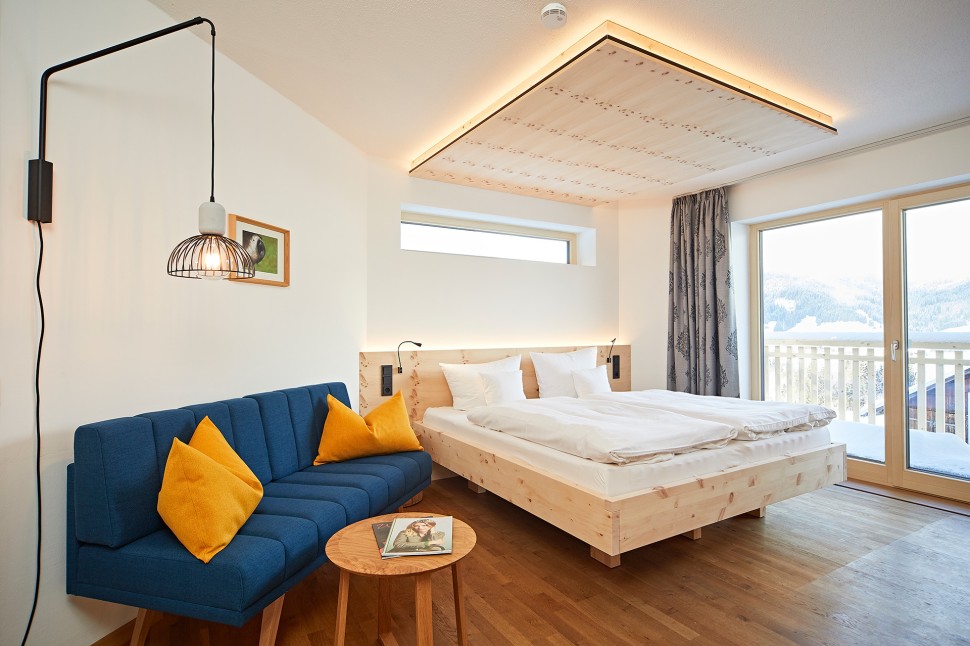 Modernes Hotelzimmer mit blauem Sofa, Beleuchtung, Fernseher und Balkon in hellen Holzfarben im Kleinwalsertal Hotel Oswalda Hus.