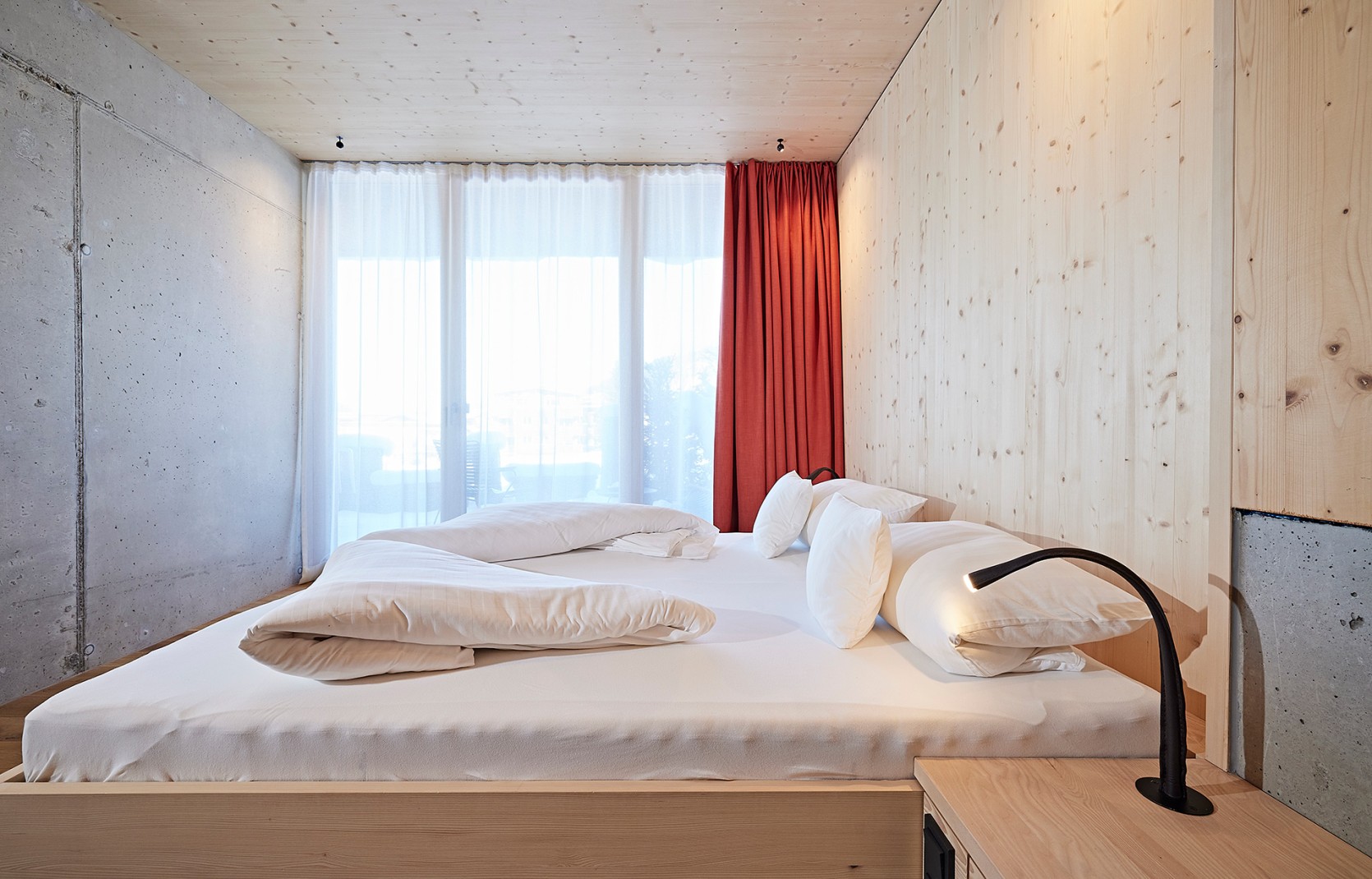 Schlichtes Hotelzimmer mit Holzwänden, Sichtbeton und roten Vorhängen im 4 Sterne Hotel Oswalda Hus im Kleinwalsertal.