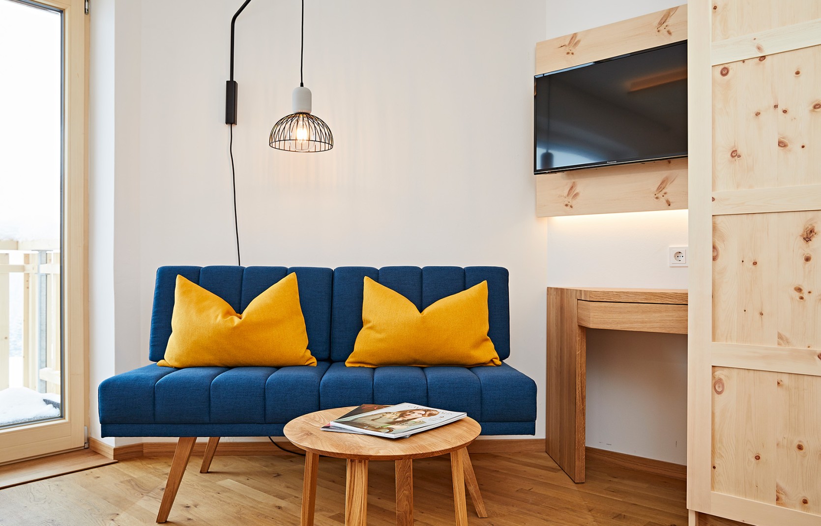 Modernes Hotelzimmer mit blauem Sofa, Beleuchtung, Fernseher und Balkon in hellen Holzfarben im Kleinwalsertal Hotel Oswalda Hus.
