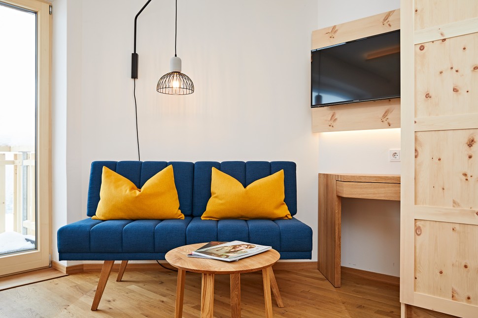 Modernes Hotelzimmer mit blauem Sofa, Beleuchtung, Fernseher und Balkon in hellen Holzfarben im Kleinwalsertal Hotel Oswalda Hus.